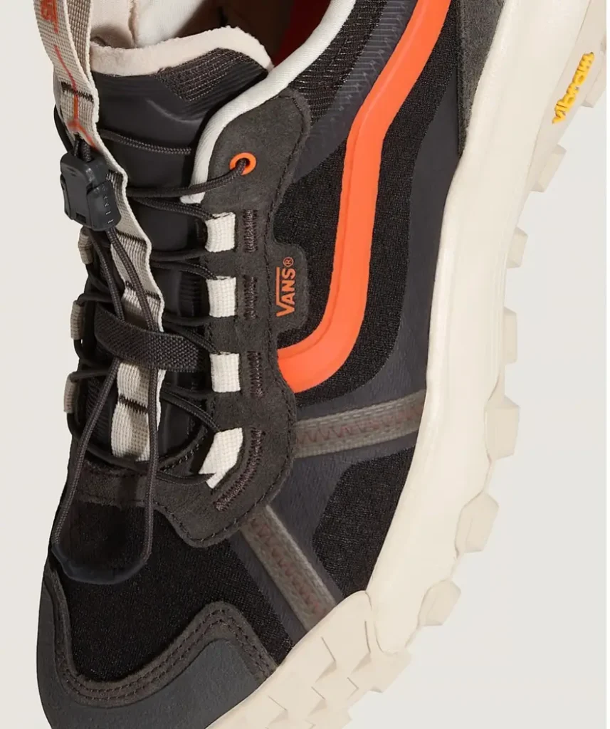 Close-up angled view of the Vans MTE Travel Pack shoe featuring a black technical mesh upper, vivid orange side stripe, toggle quick-lace system, fleece-lined collar, layered weather-resistant overlays, and chunky off-white Vibram outsole built for all-terrain travel and outdoor performance