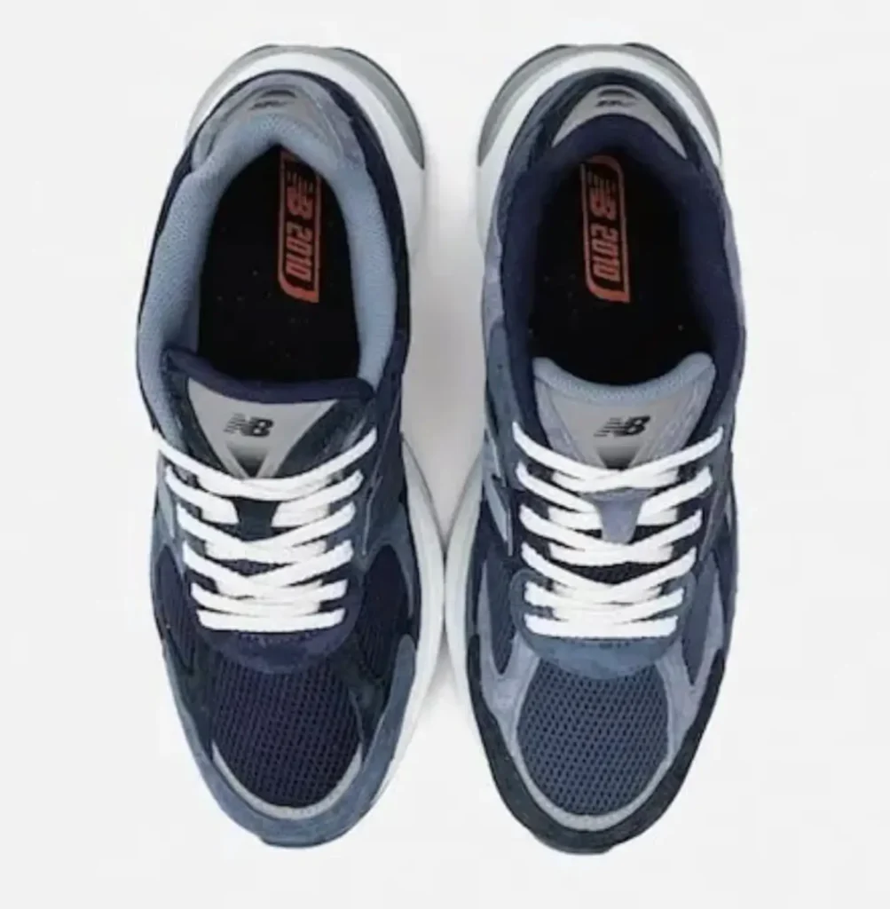 Top-down studio view of the New Balance x BEAMS 2010 “Navy” shoe highlighting asymmetrical navy paneling, breathable mesh toe boxes, white rope laces, and branded BEAMS insoles set against a clean light-grey backdrop