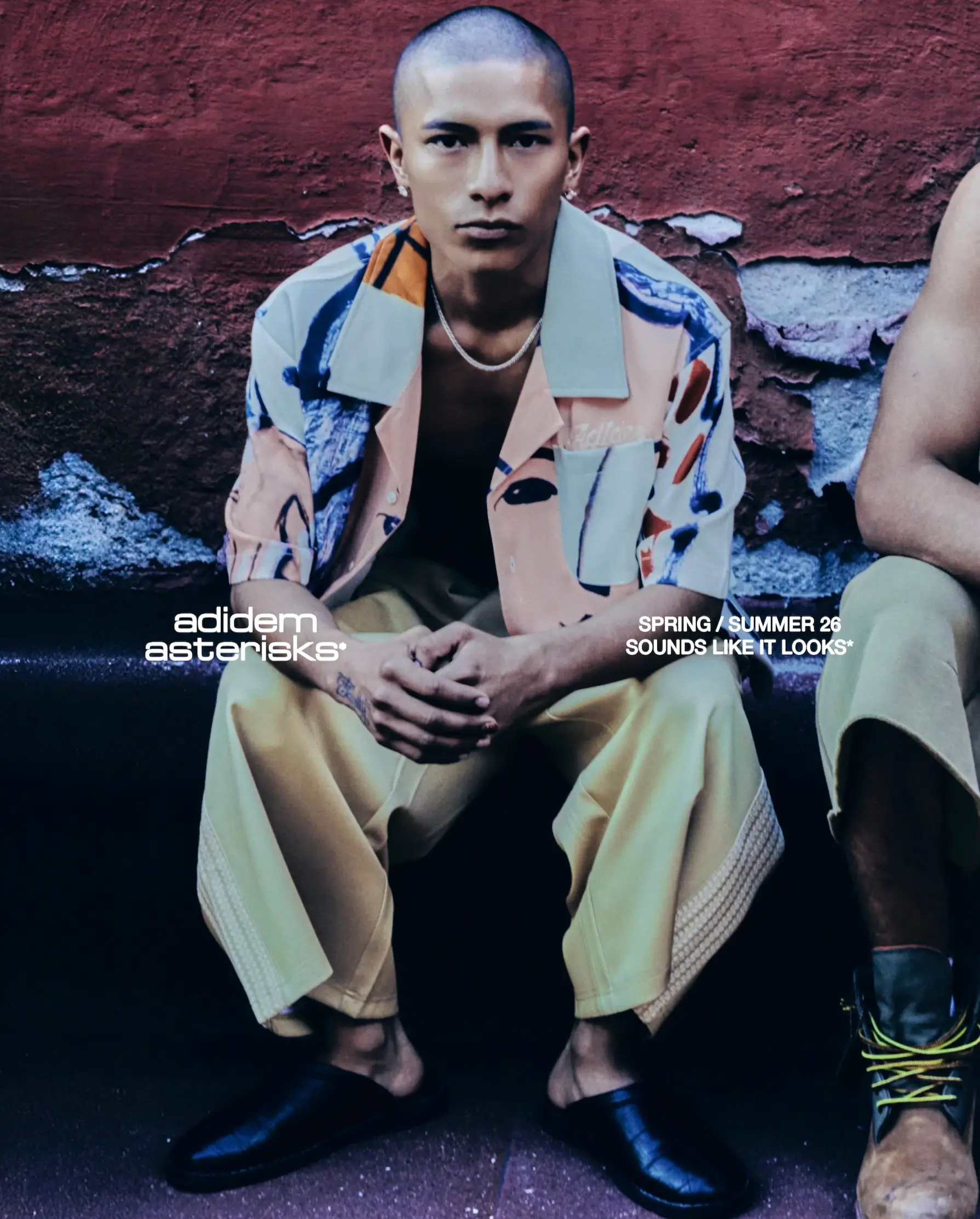 Campaign image for Adidem Astericks Spring/Summer 2026 “Sounds Like It Looks” featuring a seated model wearing the Faces print camp collar shirt in soft blush, navy, cream, and rust tones with tonal embroidered chest script, relaxed cropped trousers, and black leather slip-ons against a distressed burgundy wall backdrop