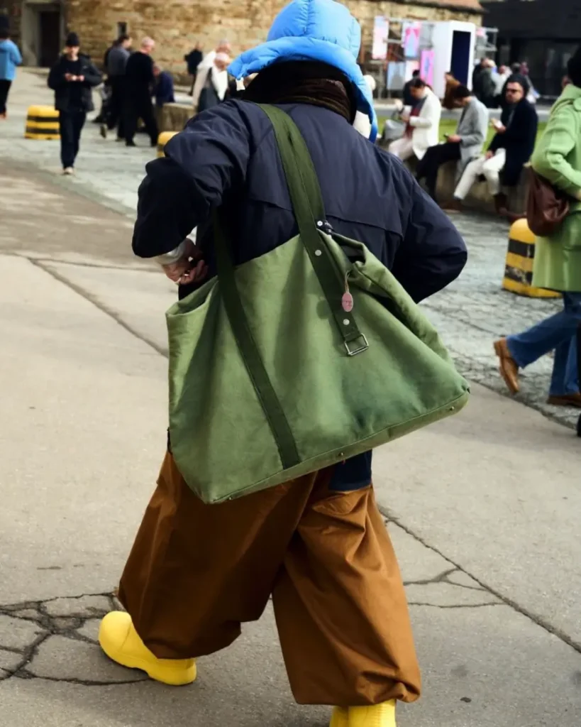 Rafaello Napoleon walking outdoors in a street-style setting wearing a bright blue padded hat, dark jacket, and wide brown trousers, carrying a large olive green canvas tote bag with a long strap and metal buckle, paired with bold yellow footwear, with people gathered in the background near a stone building