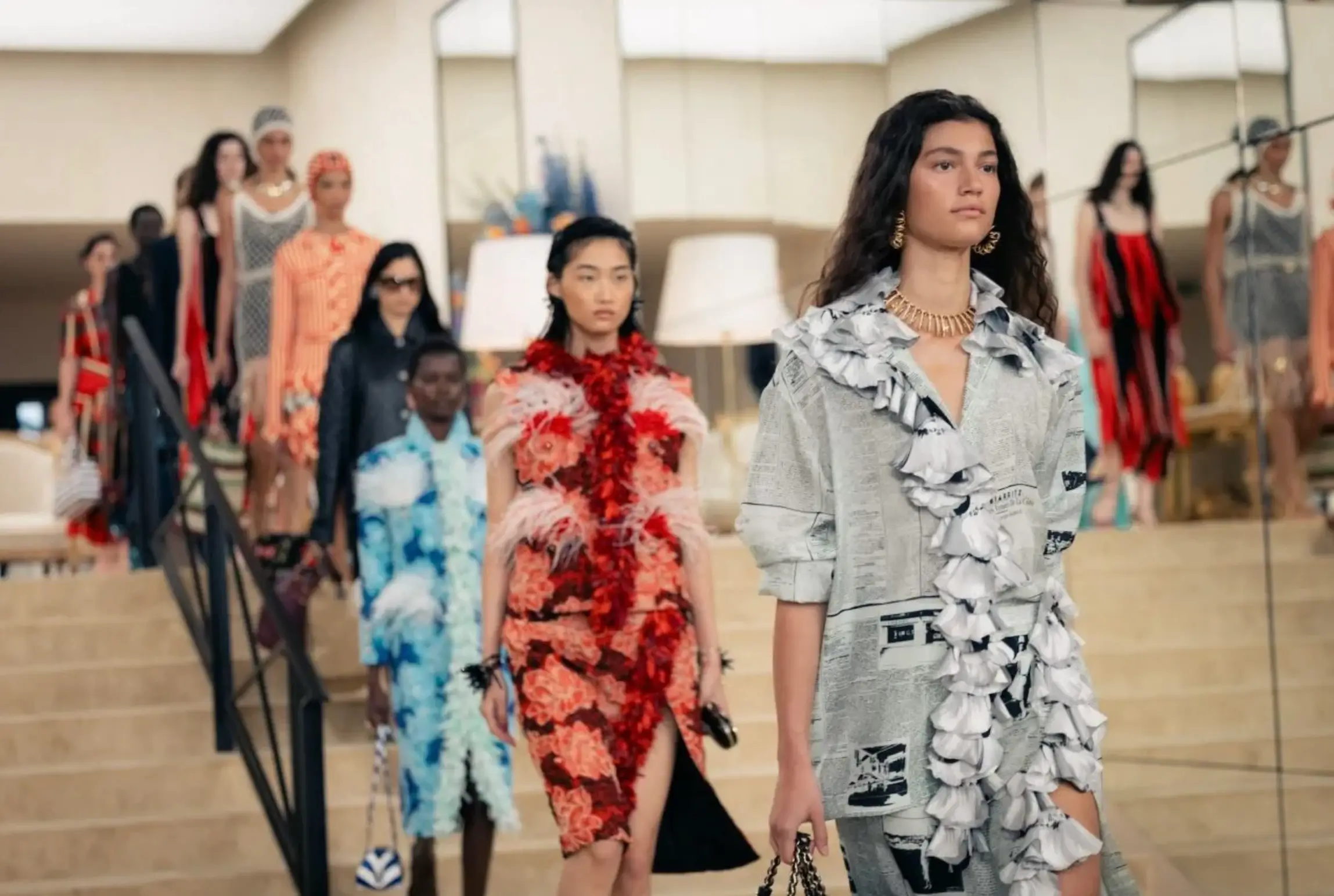 Runway models descending a staircase in a bright interior setting, led by a look featuring a structured grey ensemble with ruffled detailing and subtle graphic prints, followed by vibrant floral and textured dresses, capturing a layered, resort-inspired Chanel presentation