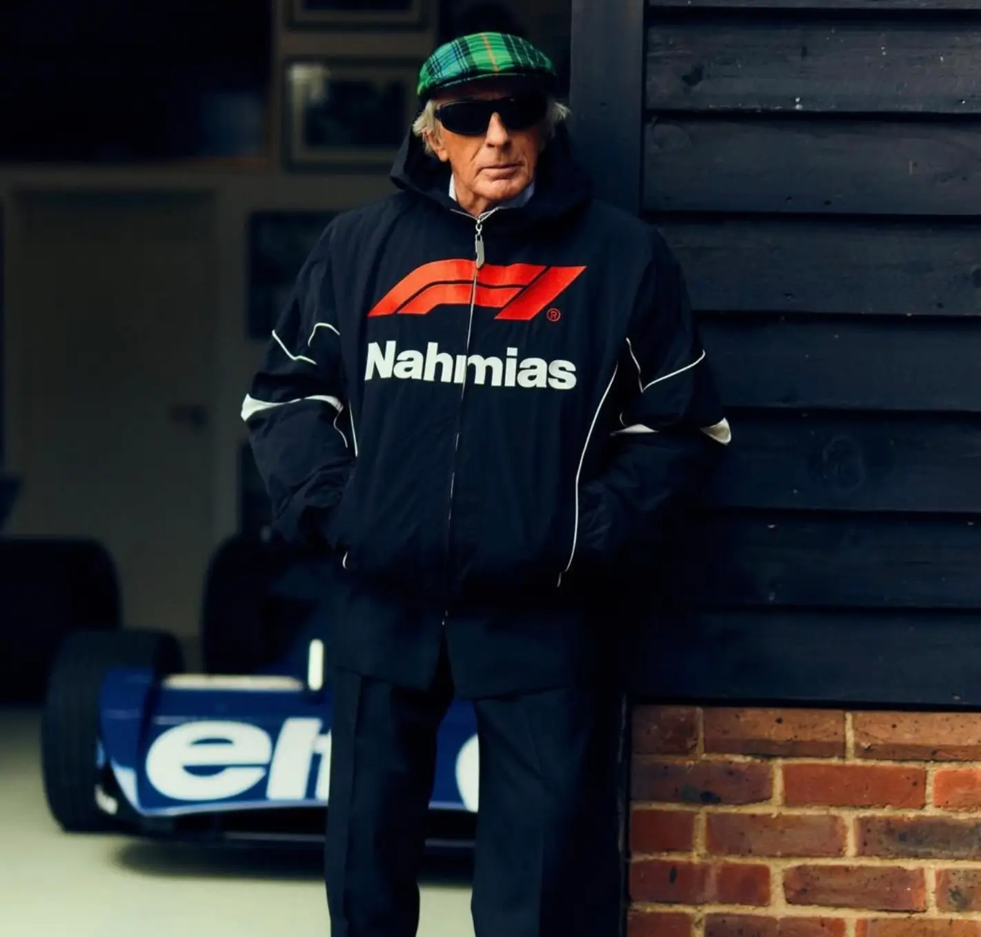 Black racing jacket worn in a lifestyle setting, featuring a bold front graphic with an oversized red Formula 1 logo above large white “Nahmias” lettering. The jacket has a relaxed, slightly cropped fit with a full zip closure, elasticated hem, and subtle white piping along the sleeves, reinforcing its motorsport DNA. The look is styled with dark trousers, a green plaid cap, and sunglasses, blending heritage elements with contemporary streetwear. Set against a garage backdrop with a Formula 1 car partially visible, the scene underscores the authentic racing influence while presenting the piece in a cinematic, real-world context