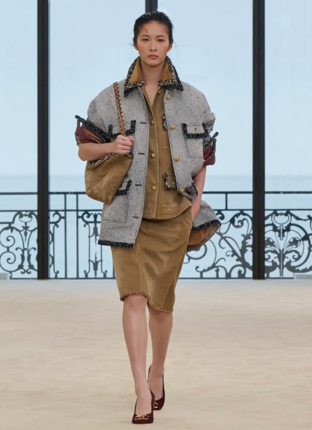 A runway look featuring layered Chanel styling with a relaxed grey tweed jacket trimmed in textured black edging over tonal camel separates, including a utility-inspired shirt and matching knee-length skirt. The look is finished with a soft shoulder bag on a chain strap and deep burgundy pumps, balancing house tailoring codes with an understated, contemporary silhouette