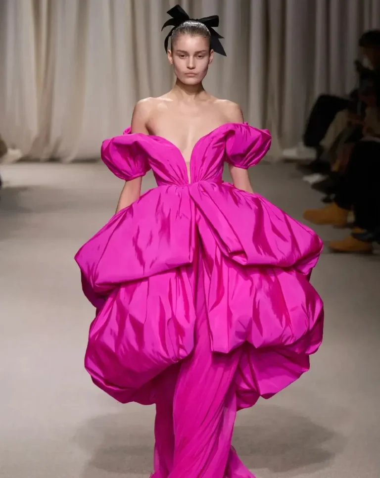 Runway model walking in an oversized fuchsia couture gown featuring dramatic sculpted volume, puff sleeves, and a plunging neckline. The glossy fabric creates exaggerated gathered forms while a black bow headpiece adds contrast against a neutral runway backdrop