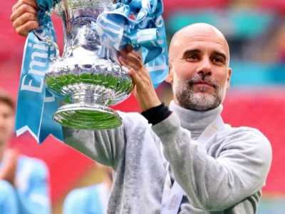 A celebratory stadium portrait of a football manager holding a large silver cup trophy decorated with pale blue ribbons while wearing a light grey turtleneck knit. A winner’s medal hangs around his neck as blurred teammates and stadium seating create a colorful background. The image captures a composed moment of triumph, balancing sporting achievement with an understated, elevated touchline style aesthetic