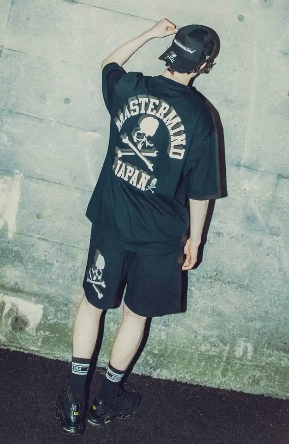 A male model wearing the New Era x mastermind JAPAN 2026 collection, viewed from the back. He is dressed in a full black outfit featuring an oversized short-sleeve t-shirt with a large arched 'MASTERMIND JAPAN' logo and skull & crossbones graphic on the back, matching black shorts with a smaller skull motif on the left leg, a black trucker cap with mastermind branding, and black sneakers. The model stands against a concrete wall with one arm raised, adjusting his cap