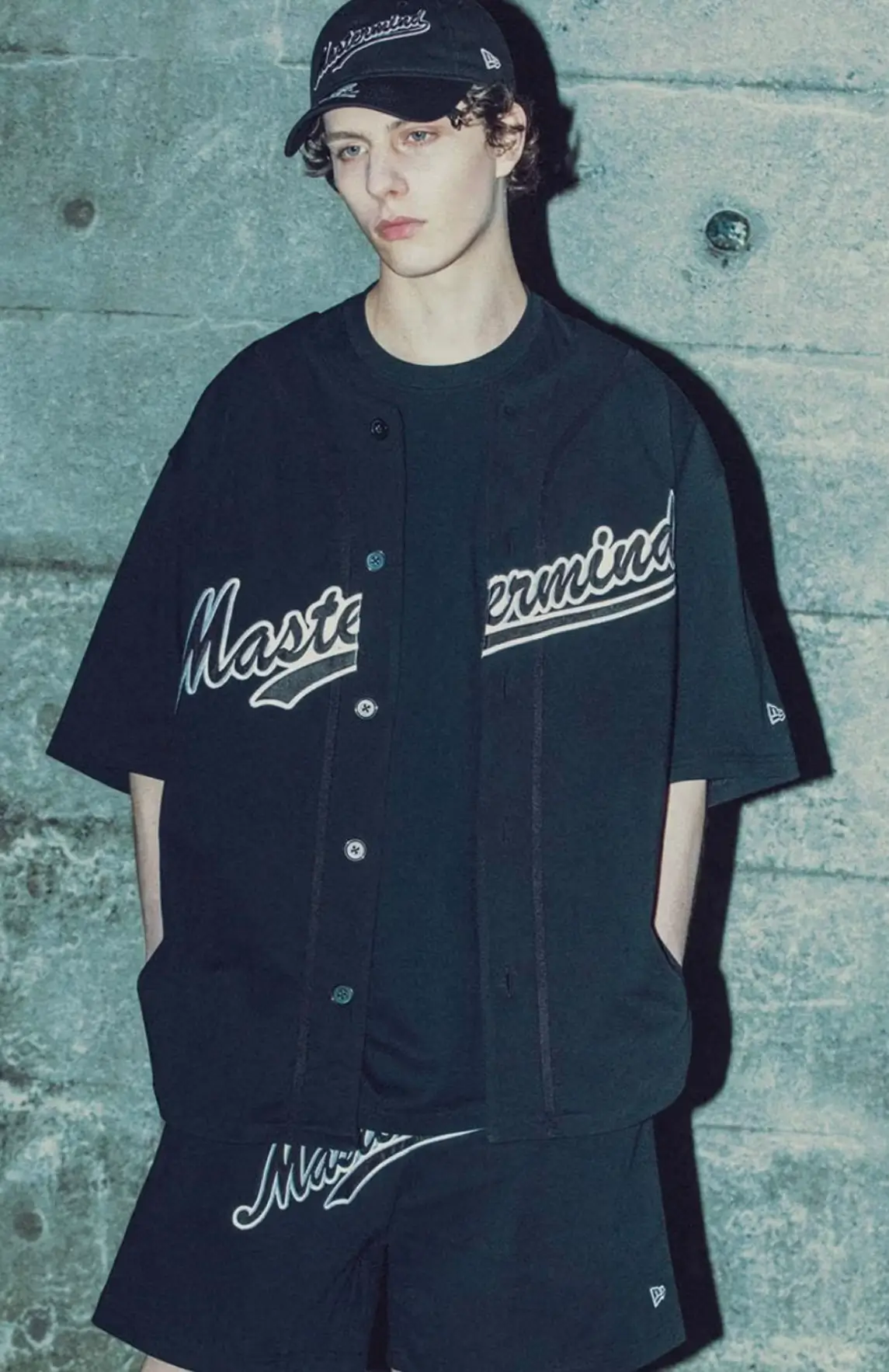 A male model wearing the New Era x mastermind JAPAN 2026 collection, standing against a concrete wall and looking directly at the camera. He is styled in a full black outfit: an oversized button-up baseball jersey with 'Mastermind' script across the chest, matching black shorts with 'Mastermind' script on the leg, and a black New Era trucker cap with 'mastermind' script and skull logo. His hands are in his pockets, showcasing the complete coordinated set