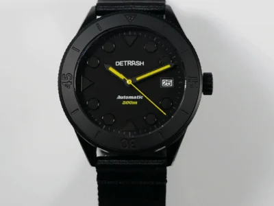 Front-facing product shot of the Detrash Gotham’s Vigilante 2 automatic dive watch featuring a monochrome stealth-black case and dial with bright yellow hands and accents. Circular and triangular hour markers create a minimalist tactical look, while the matte dive bezel, date window at 3 o’clock, and black recycled strap reinforce the watch’s Batman-inspired aesthetic and understated utilitarian design
