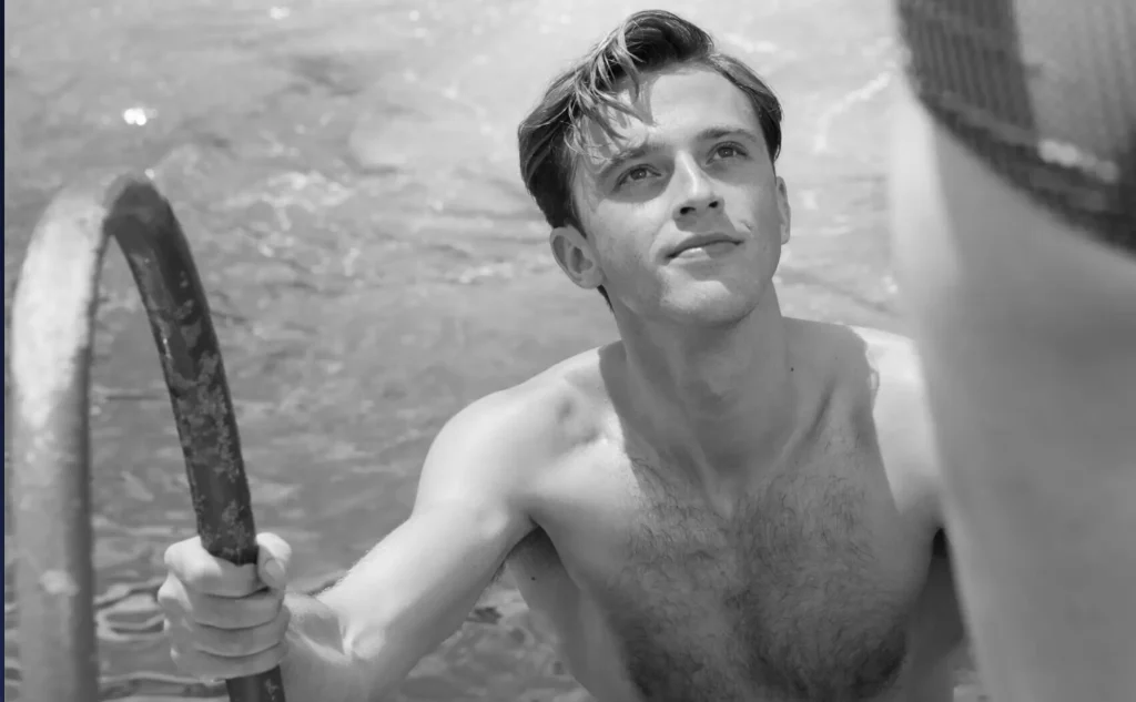 Black-and-white film still showing a young man emerging from the water and gripping a metal pool ladder while looking upward with a calm, attentive expression. Sunlight reflects across the water and his face, creating a quiet yet intimate moment that emphasizes the film’s sensual, heat-soaked atmosphere and emotional ambiguity