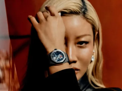 Close-up editorial portrait of a model with platinum blonde hair posing against a deep red backdrop while wearing a dark titanium-toned luxury sports watch with an openworked dial and vivid red tourbillon detail. The model partially covers one eye with her hand, emphasizing the sculptural case shape and contemporary high-fashion style