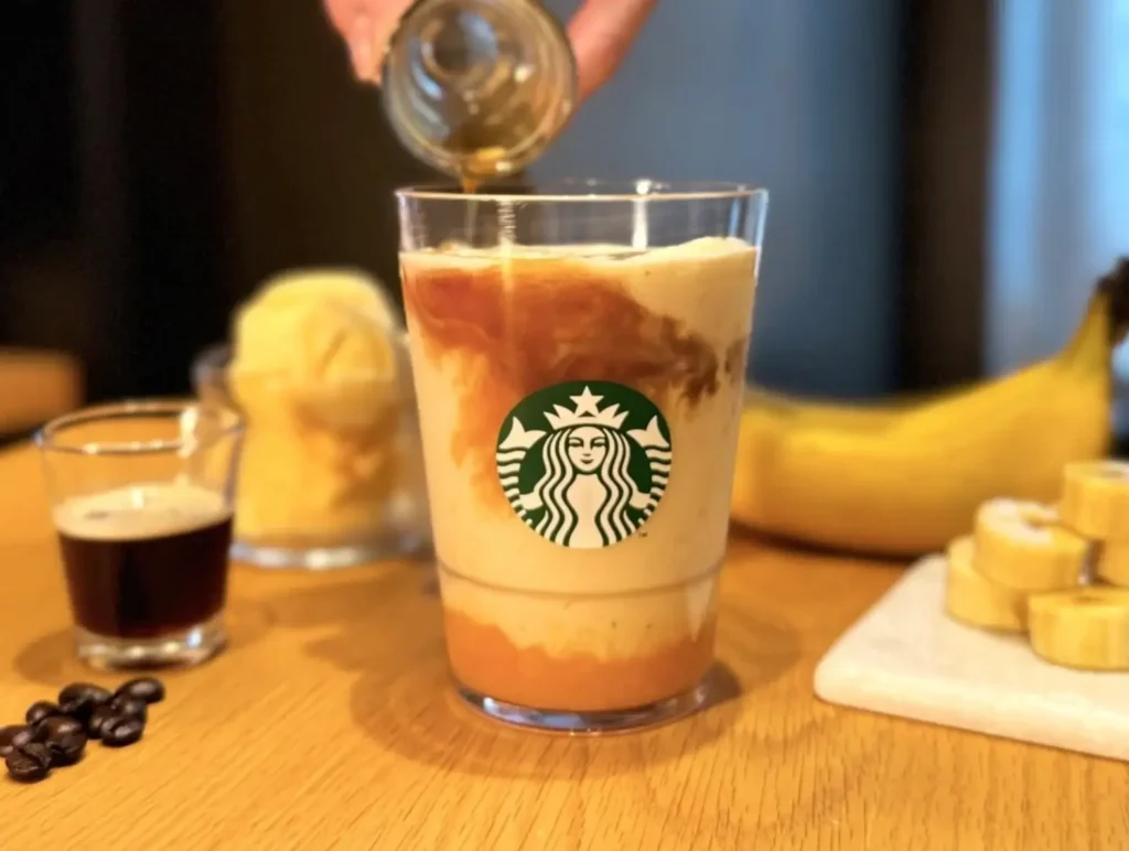 Banana Affogato Frappuccino in a Starbucks cup as espresso is poured over a creamy banana blend, creating layered swirls above real banana pulp with coffee beans and fresh banana in the background