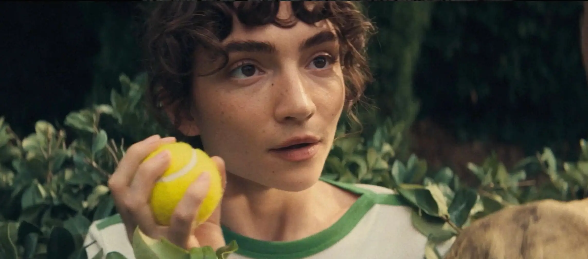 Close-up of a woman with curly hair and soft, natural makeup, holding a bright yellow tennis ball near her face. She is partially framed by green foliage, wearing a white top with green trim, her expression attentive and slightly surprised as she looks off-camera