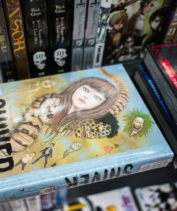 Close-up bookshelf scene featuring a colorful hardcover collection of Junji Ito’s work displayed among manga volumes and graphic novels. The illustrated cover combines surreal horror imagery—distorted faces, grotesque creatures, and dreamlike symbolism—with soft pastel tones, reflecting Ito’s signature balance between beauty, absurdity, and psychological dread within contemporary horror manga culture