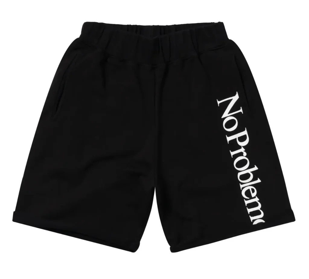 Black relaxed-fit shorts featuring oversized white “No Problemo” typography running down one leg, styled with an elastic waistband and minimalist studio presentation against a light-gray background