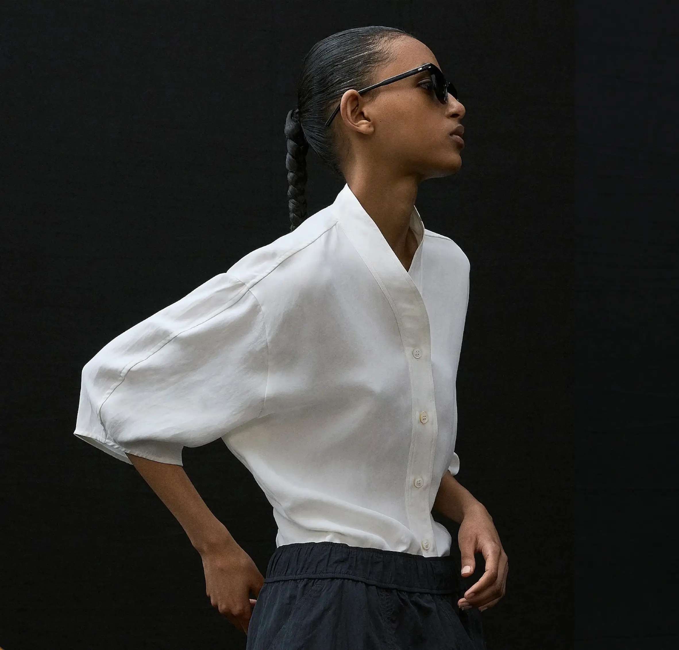 Model wearing a sculptural white short-sleeve shirt tucked into relaxed black technical trousers with black sunglasses and a braided hairstyle, photographed against a dark studio backdrop