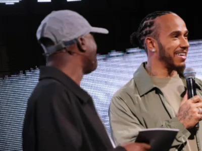 Lewis Hamilton smiles while speaking into a microphone during a lululemon event appearance, standing beside another presenter onstage against a large illuminated digital backdrop