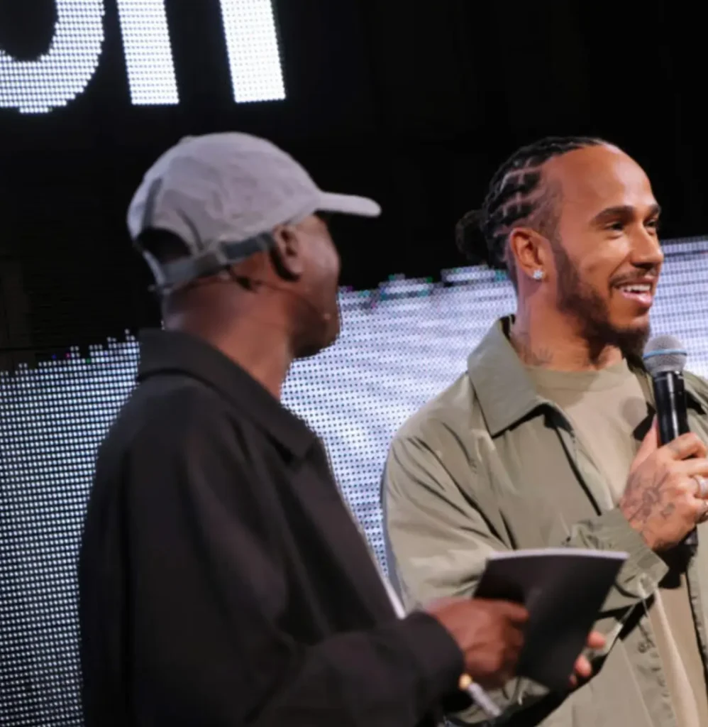 Lewis Hamilton smiles while speaking into a microphone during a lululemon event appearance, standing beside another presenter onstage against a large illuminated digital backdrop