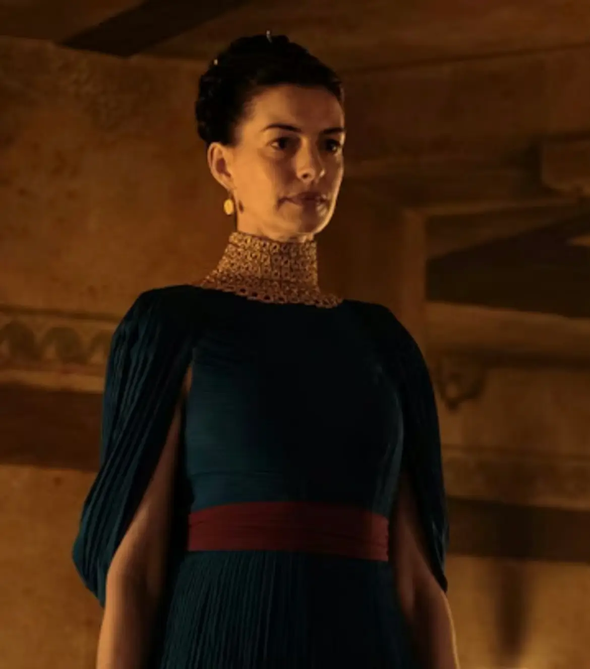A dramatic cinematic portrait of a woman standing within a warm torchlit stone interior, dressed in an elegant dark pleated gown with sculptural draped sleeves and a deep crimson waist accent. The look is elevated by an ornate gold high-neck collar piece and delicate drop earrings, blending ancient regal styling with modern couture minimalism. Her composed expression and upright posture project authority and restraint, while the amber lighting casts soft shadows across the architectural backdrop, creating an atmosphere reminiscent of mythological epics and prestige historical cinema