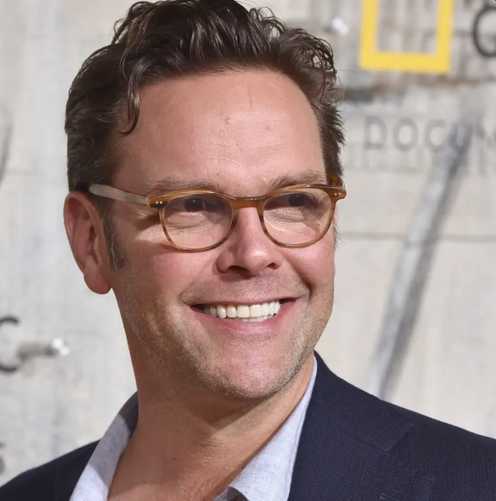 Close-up portrait of a media executive smiling during a public event appearance, wearing translucent amber eyeglasses, a navy blazer, and a light open-collar shirt against a softly blurred branded backdrop