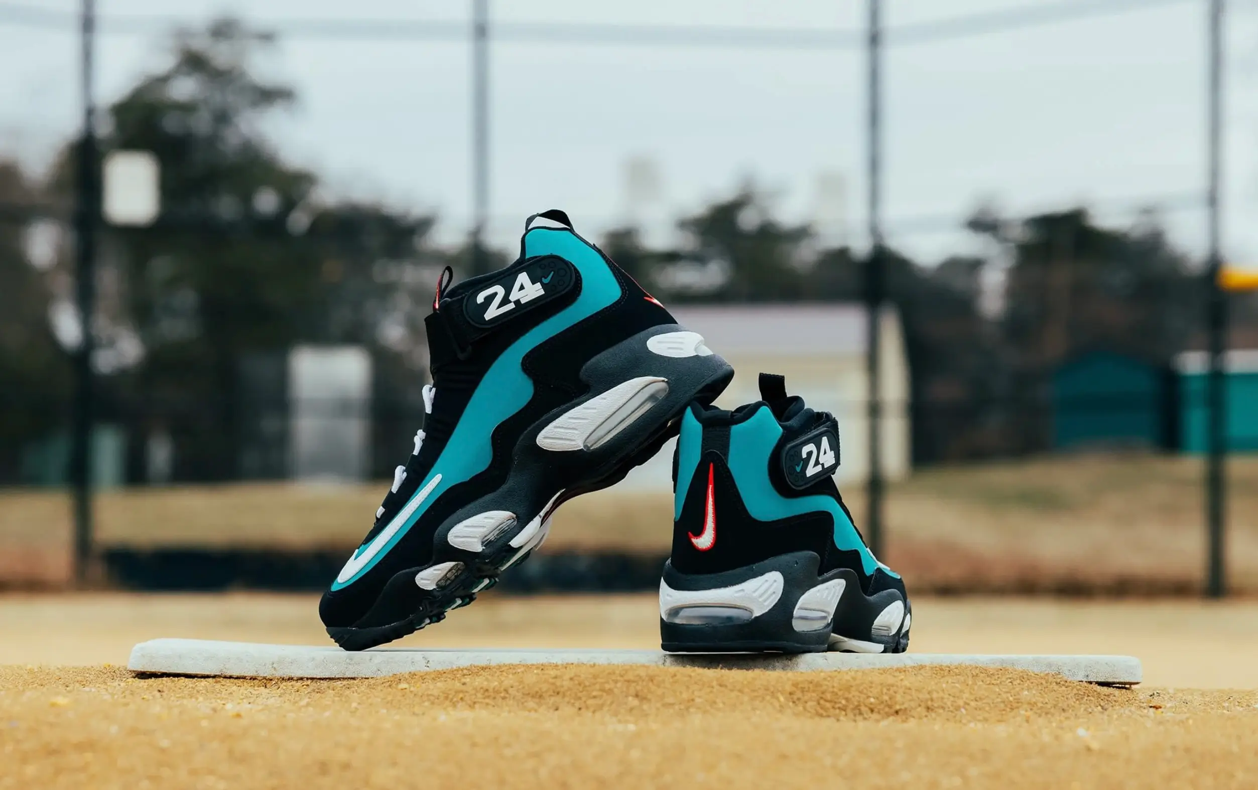 Pair of black-and-teal Nike Griffey-inspired high-top shoe posed on a baseball field base, featuring flowing teal overlays, visible Air cushioning units, and velcro ankle straps marked with the number “24.” One shoe leans diagonally against the other, highlighting the oversized white outlined Swoosh and red embroidered heel Swoosh details. The blurred outdoor baseball diamond and fencing in the background reinforce the silhouette’s retro baseball heritage aesthetic