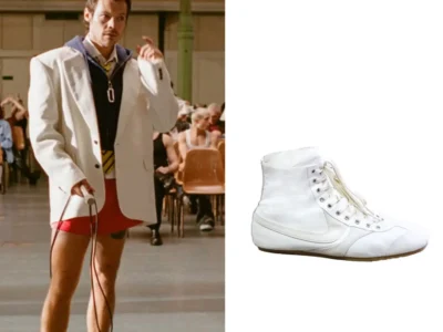 Harry Styles appears mid-performance inside the warm-toned gymnasium setting of the “Dance No More” music video, wearing an oversized ivory blazer layered over a navy zip-up hoodie and striped tie, contrasted sharply by ultra-short custom red Marc Jacobs athletic shorts. The image emphasizes the tension between tailored sophistication and provocative sportswear minimalism, while a side-by-side product cutout highlights the sleek white high-top dance-style footwear grounding the look in retro athletic elegance