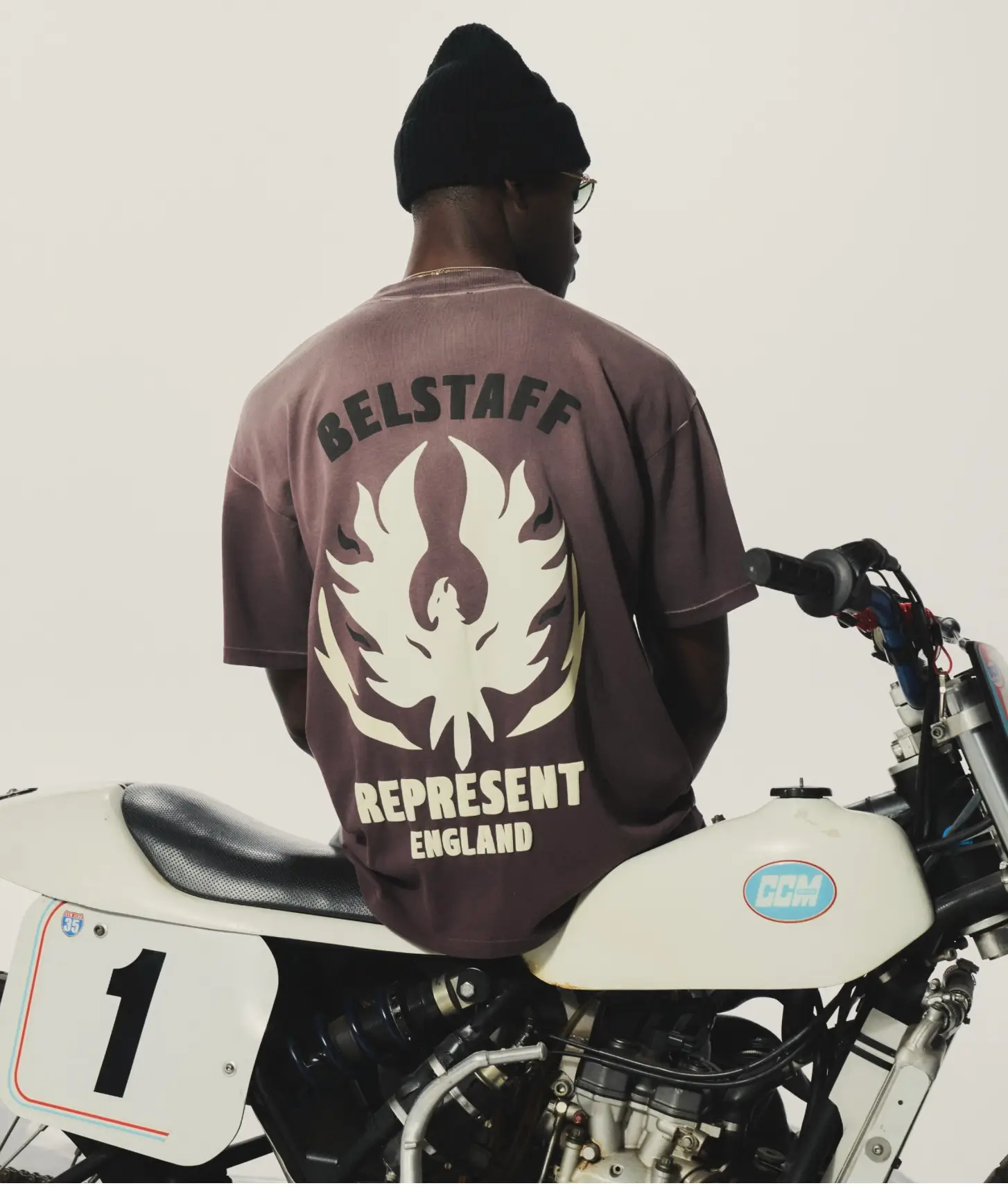 Model wearing an oversized washed plum graphic T-shirt featuring bold Belstaff x Represent phoenix branding sits on a vintage white racing motorcycle, with minimalist studio lighting emphasizing the collaboration’s fusion of British moto heritage, heavyweight streetwear graphics, and contemporary oversized styling