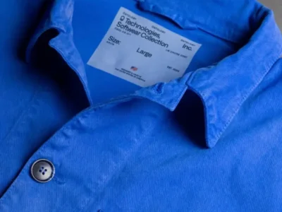 Close-up product shot of Palantir Technologies’ blue chore coat laid flat against a dark background, highlighting the structured collar, tonal stitching, matte button closure, and interior label reading “Technologies Software Collection Inc.” The vivid workwear-inspired cotton fabric emphasizes the jacket’s minimalist industrial aesthetic and Made-in-America positioning