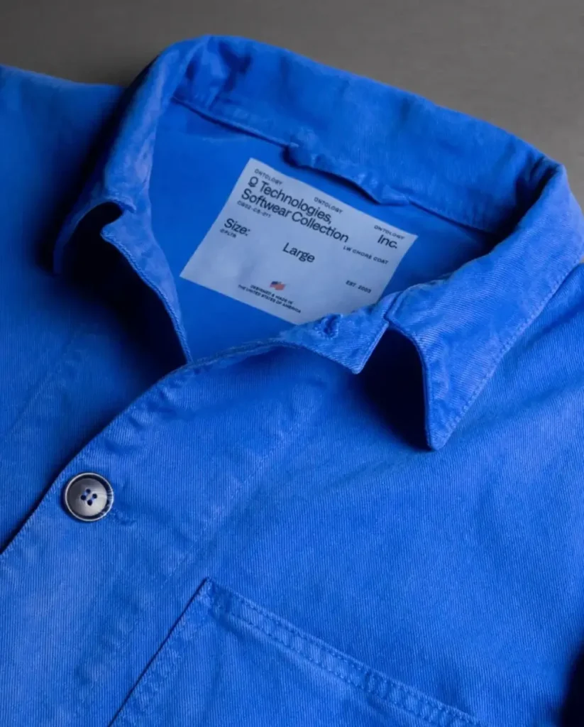 Close-up product shot of Palantir Technologies’ blue chore coat laid flat against a dark background, highlighting the structured collar, tonal stitching, matte button closure, and interior label reading “Technologies Software Collection Inc.” The vivid workwear-inspired cotton fabric emphasizes the jacket’s minimalist industrial aesthetic and Made-in-America positioning