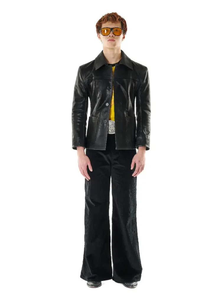 A model wears a sleek black leather jacket from the LỰU ĐẠN x Bruce Lee capsule collection, styled with flared black trousers and amber-tinted oversized sunglasses. A vivid yellow layer beneath the jacket subtly references Bruce Lee’s legendary Game of Death jumpsuit, while the visible silver statement belt buckle reinforces the collaboration’s martial arts iconography. The sharply tailored silhouette merges retro kung fu cinema, 1970s glam rebellion, and contemporary luxury streetwear into a look that feels both cinematic and confrontational