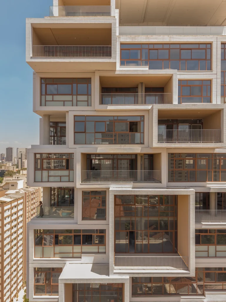 A close-up view of a contemporary residential tower reveals an intricate arrangement of stacked concrete volumes, recessed terraces, and expansive bronze-framed windows. The modular façade creates a layered geometric rhythm of balconies, cantilevered living spaces, and open-air voids, emphasizing the building’s sculptural approach to luxury urban architecture under bright midday light