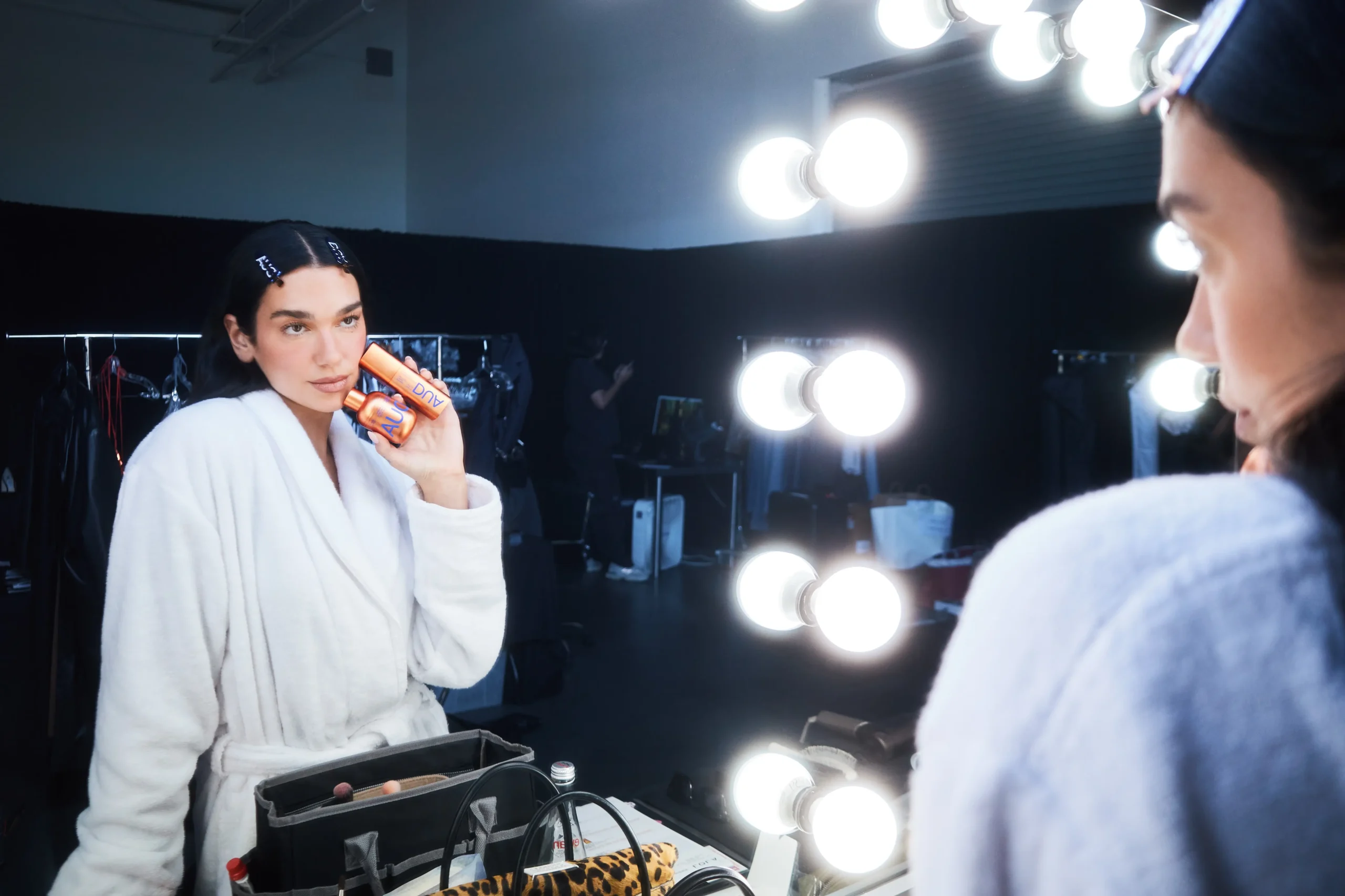 Behind-the-scenes beauty campaign image showing a woman in a white robe seated at a lighted vanity while holding DUA skincare products beside her face. Reflected in the mirror and surrounded by backstage styling equipment, the scene conveys an effortless pre-performance skincare ritual and polished modern glamour