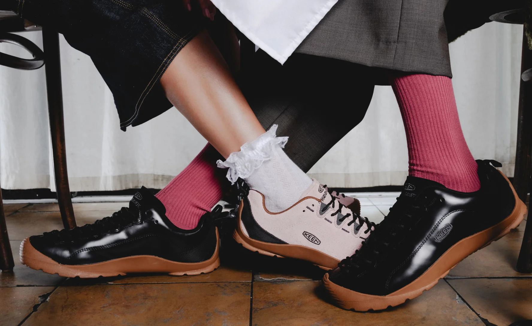 Lifestyle campaign image featuring multiple KEEN Jasper silhouettes styled in a fashion-forward indoor setting, including glossy black leather low-top pairs with gum soles and a contrasting cream-and-black hiking-inspired variation. Ribbed magenta socks, lace-trim white socks, and layered tailored garments reinforce the blend of gorpcore utility and contemporary editorial styling