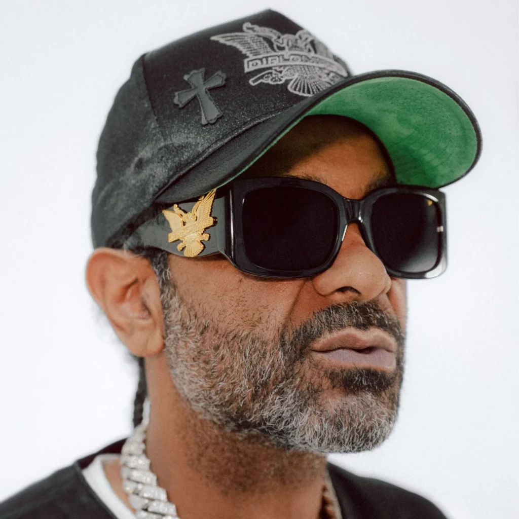 Close-up portrait of a Jim Jones hip-hop artist wearing oversized black rectangular sunglasses from the ESSX x Diplomats Vintage Frames collaboration. The glossy black frames feature a gold eagle logo emblem on the temple, paired with a black leather cap, diamond chain, and a salt-and-pepper beard against a clean studio backdro