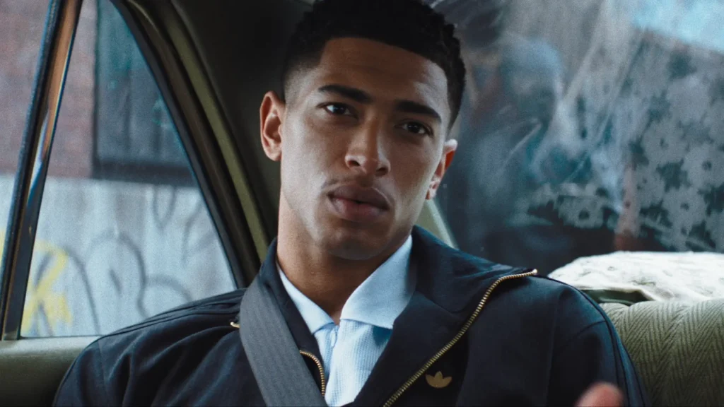 A young man sits inside the back seat of a car in a cinematic close-up portrait, wearing a dark adidas track jacket layered over a crisp white collared knit shirt. Soft natural light filters through the window beside him, highlighting his calm expression and sharply detailed haircut against the muted interior of the vehicle. Gold zipper accents and a subtle adidas Trefoil logo add understated luxury to the sporty styling, while blurred city textures outside the window create a reflective, contemporary atmosphere rooted in modern football culture and elevated streetwear aesthetics