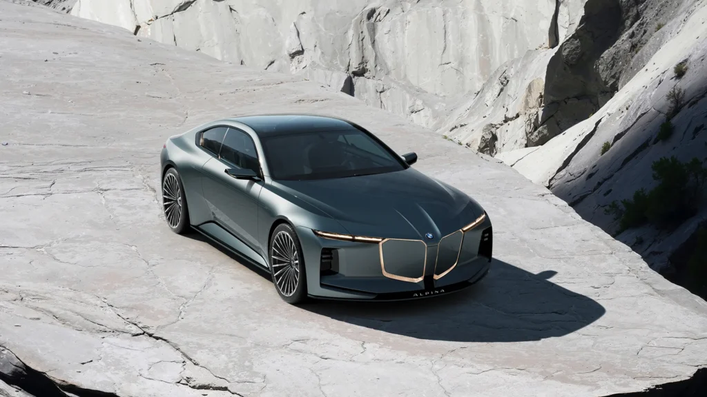 The Vision BMW ALPINA appears in full form against a stark quarry landscape, its elongated grand touring proportions and restrained surfacing amplified by the surrounding monochromatic stone. Finished in a muted metallic green-gray tone, the concept combines a dramatically low roofline with an illuminated reinterpretation of BMW’s shark-nose grille architecture, outlined by warm ambient lighting that contrasts against the cold mineral terrain. Massive multi-spoke ALPINA wheels, ultra-slim lighting signatures, and the coupe’s flowing shoulder line reinforce its balance of elegance and authority, presenting the car less as an aggressive performance machine and more as a sculptural expression of modern long-distance luxury