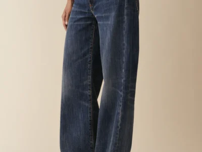Studio image of wide straight-leg Jeanerica denim jeans in a dark indigo wash with subtle faded vertical texture detailing. The low-rise rigid denim silhouette features relaxed proportions through the leg, classic five-pocket construction, and soft whiskering effects, styled with a minimal white top and pointed shoes against a neutral beige backdrop