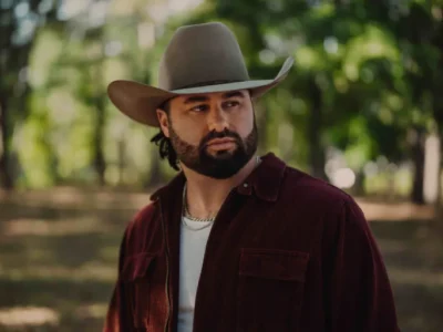 Portrait of Koe Wetzel standing outdoors in a wooded setting, wearing a beige cowboy hat, burgundy overshirt, white tank top, and silver chain while looking off-camera in warm natural light