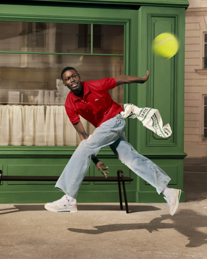 A man in a red polo shirt and light blue jeans is captured mid-stride in a dynamic leap, reaching toward a bright yellow tennis ball suspended in the air. He wears white sneakers and moves in front of a green storefront with large windows and soft curtains, a bench behind him adding to the street-scene setting. A white scarf with green accents trails from his hand, echoing the motion and energy of the moment