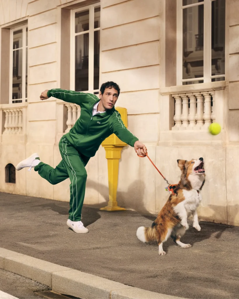 A man in a green tracksuit lunges forward mid-step on a city sidewalk, holding a leash as a small brown-and-white dog jumps up excitedly toward a bright yellow tennis ball in midair. Both appear animated and focused on the ball, set against a cream-colored building with tall windows and a vivid yellow post nearby, capturing a playful, energetic street moment