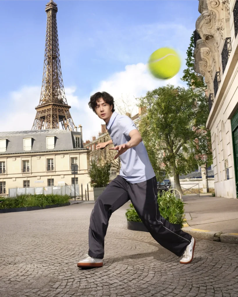 A man stands in a wide, grounded stance on a cobblestone street, reaching forward toward a bright yellow tennis ball flying through the air. He wears a light blue polo shirt, dark loose-fitting pants, and white sneakers with gum soles. Behind him, the Eiffel Tower rises prominently against a blue sky, framed by classic Parisian buildings and greenery, creating a dynamic moment that blends urban movement with an iconic landmark backdrop