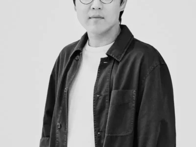 Portrait of Jongjin Park wearing round glasses and a dark overshirt layered over a white T-shirt, photographed in a minimalist black-and-white studio setting following his 2026 LOEWE FOUNDATION Craft Prize win for Strata of Illusion