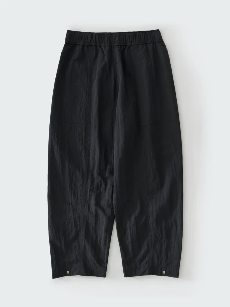 Black technical nylon trousers with a voluminous tapered silhouette, elastic waistband, curved seam construction, and subtle hem snap details photographed against a pale gray background