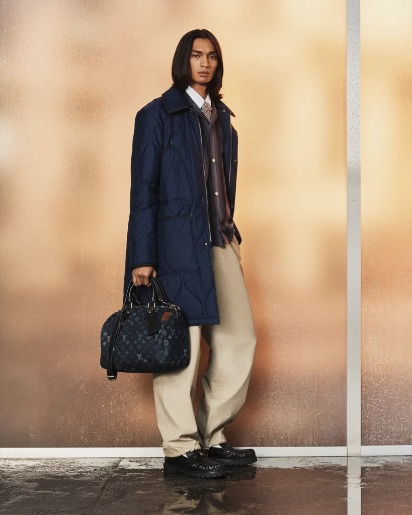 Tailored navy quilted coat layered over shirt-and-tie styling with relaxed trousers and a monogram Louis Vuitton duffel