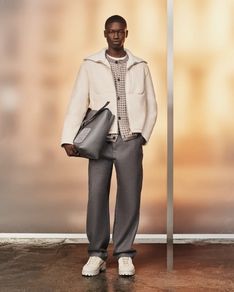 Model styled in an ivory zip-front jacket, patterned knit cardigan, gray trousers, and white sneakers while carrying a soft gray leather bag