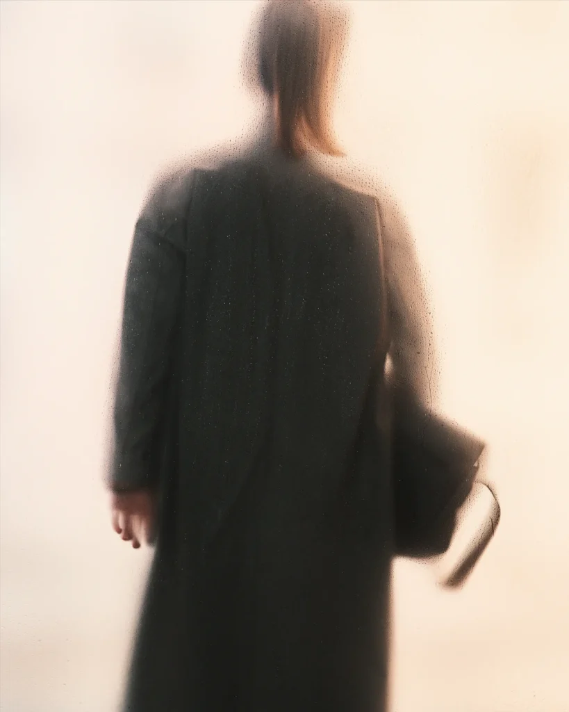 Blurred silhouette behind frosted glass highlighting the elongated shape of a tailored overcoat and leather carryall
