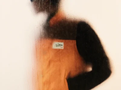 Dreamlike rear-view silhouette behind misted glass showcasing a mustard-yellow hooded Louis Vuitton coat and monogram travel bag