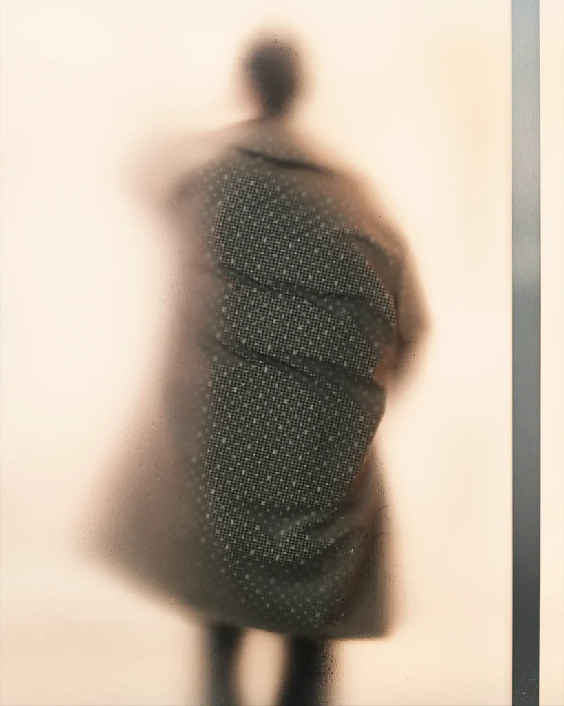 Blurred silhouette behind frosted glass highlighting the elongated shape of a tailored overcoat and leather carryall