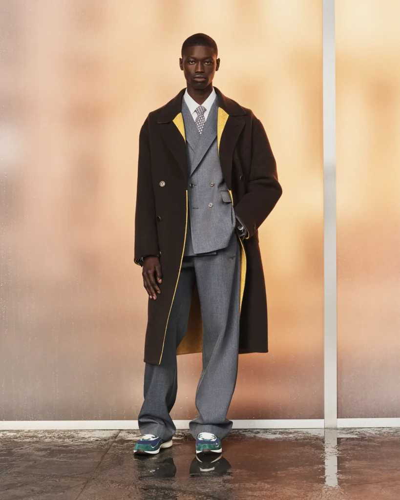 Model wearing a charcoal overcoat layered over a gray double-breasted suit with yellow monogram lining accents and athletic-inspired footwear