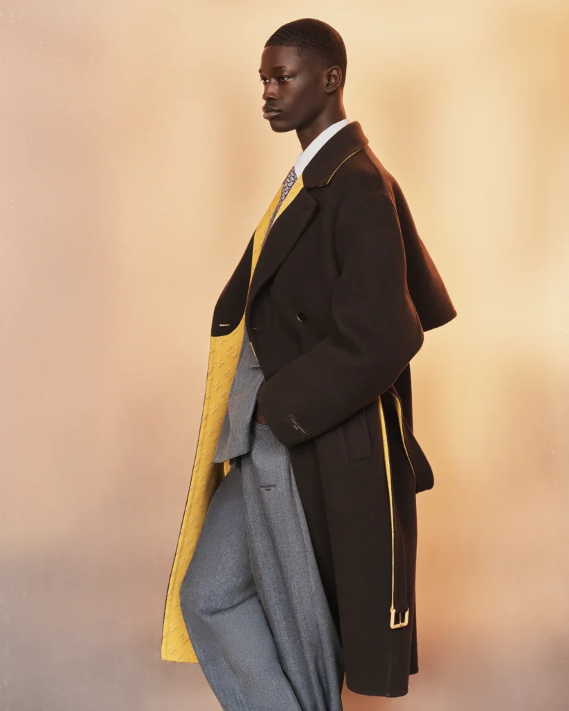 Side profile highlighting the flowing silhouette of a dark overcoat with yellow monogram interior layered over gray tailoring