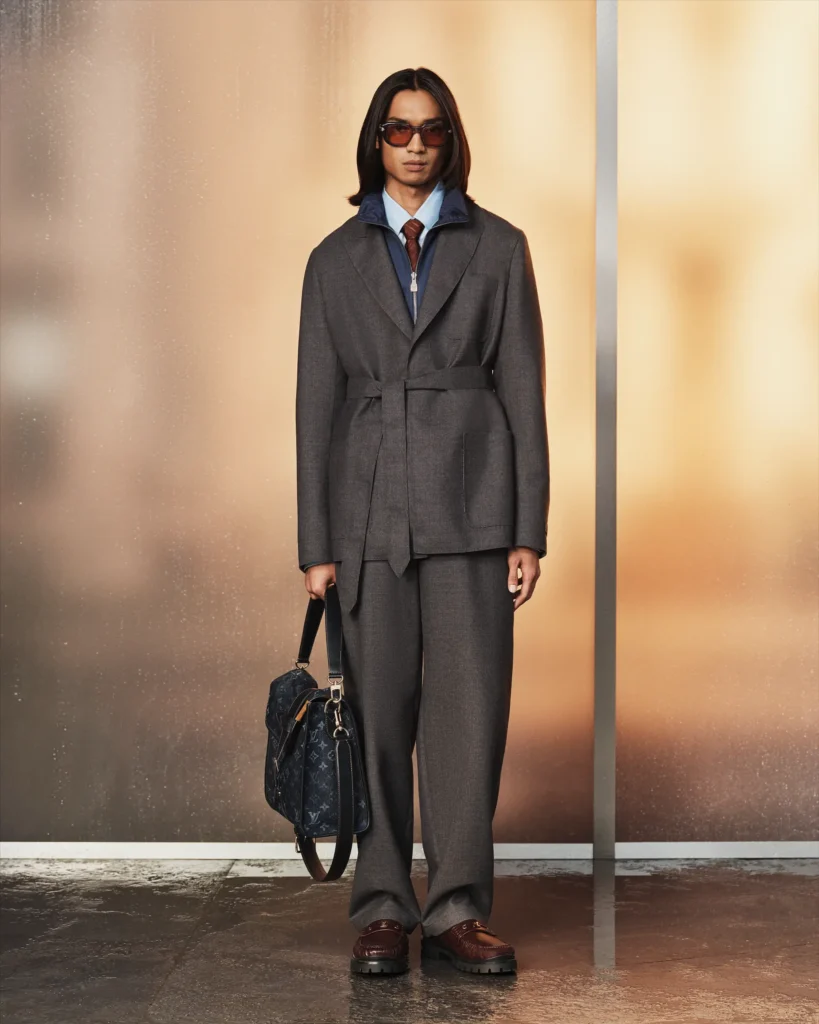 Model in a gray belted suit with a navy zip-neck layer, burgundy tie, sunglasses, and a Louis Vuitton monogram briefcase